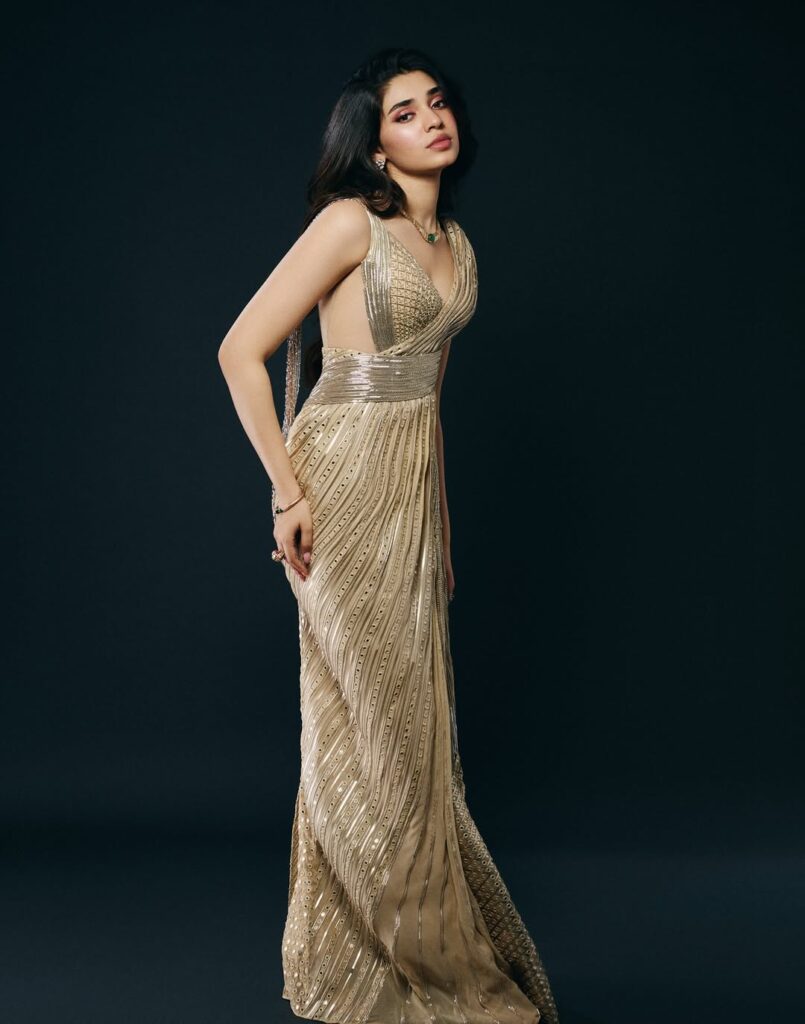 Krithi Shetty wearing an elegant golden gown, posing gracefully in a studio photoshoot with a dark background, showcasing a stylish and glamorous look.