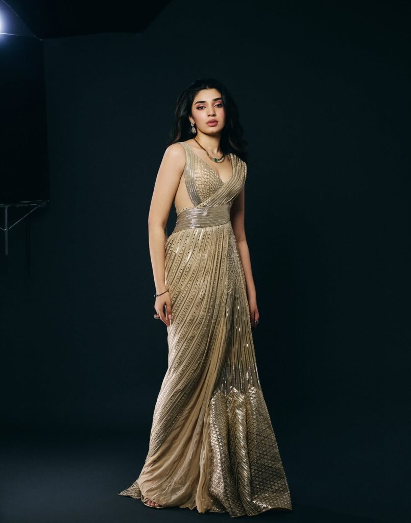 Krithi Shetty wearing an elegant golden gown, posing gracefully in a studio photoshoot with a dark background, showcasing a stylish and glamorous look.