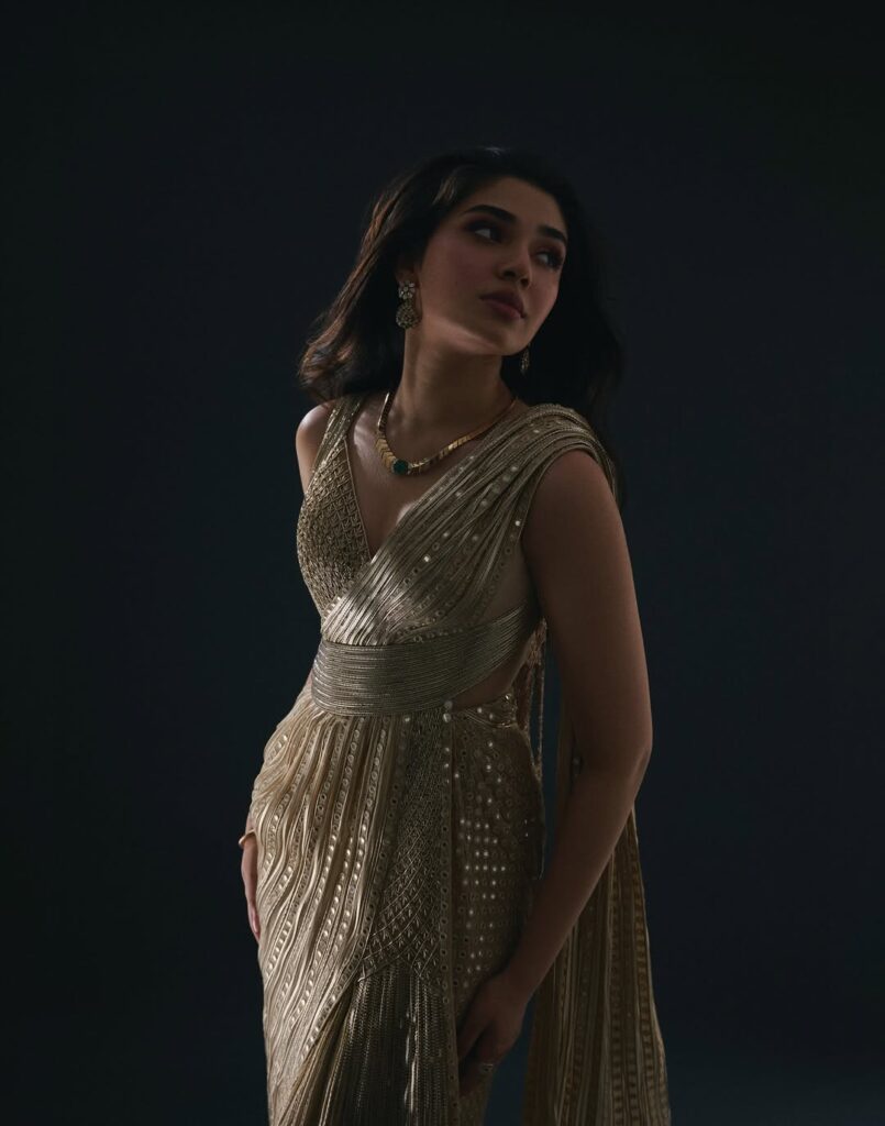 Krithi Shetty wearing an elegant golden gown, posing gracefully in a studio photoshoot with a dark background, showcasing a stylish and glamorous look.