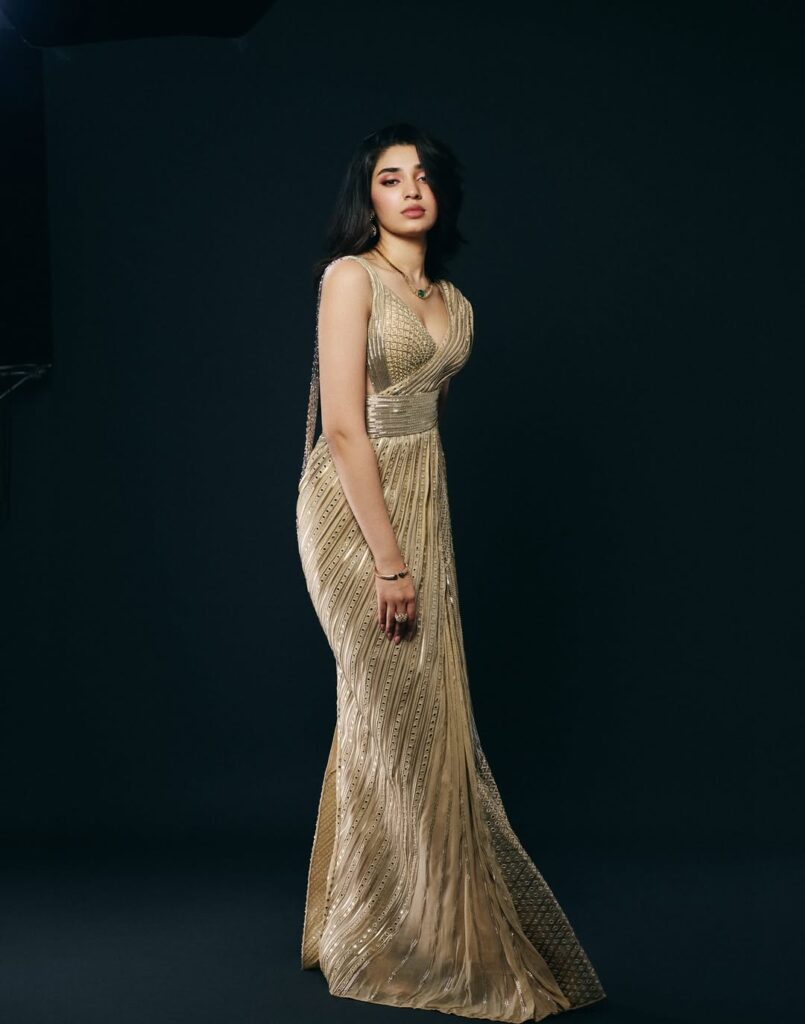 Krithi Shetty wearing an elegant golden gown, posing gracefully in a studio photoshoot with a dark background, showcasing a stylish and glamorous look.